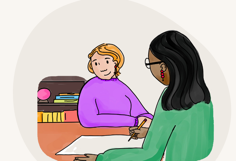 A parent and teacher sit together in a classroom setting. The teacher is taking notes.